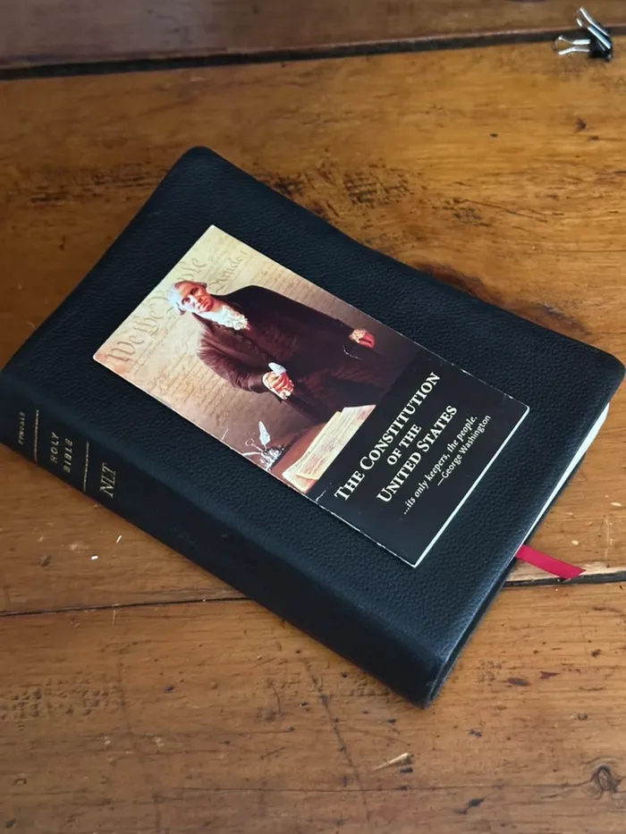A black leather Bible on a wooden desk with a bookmark of the Constitution of the United States resting on top.