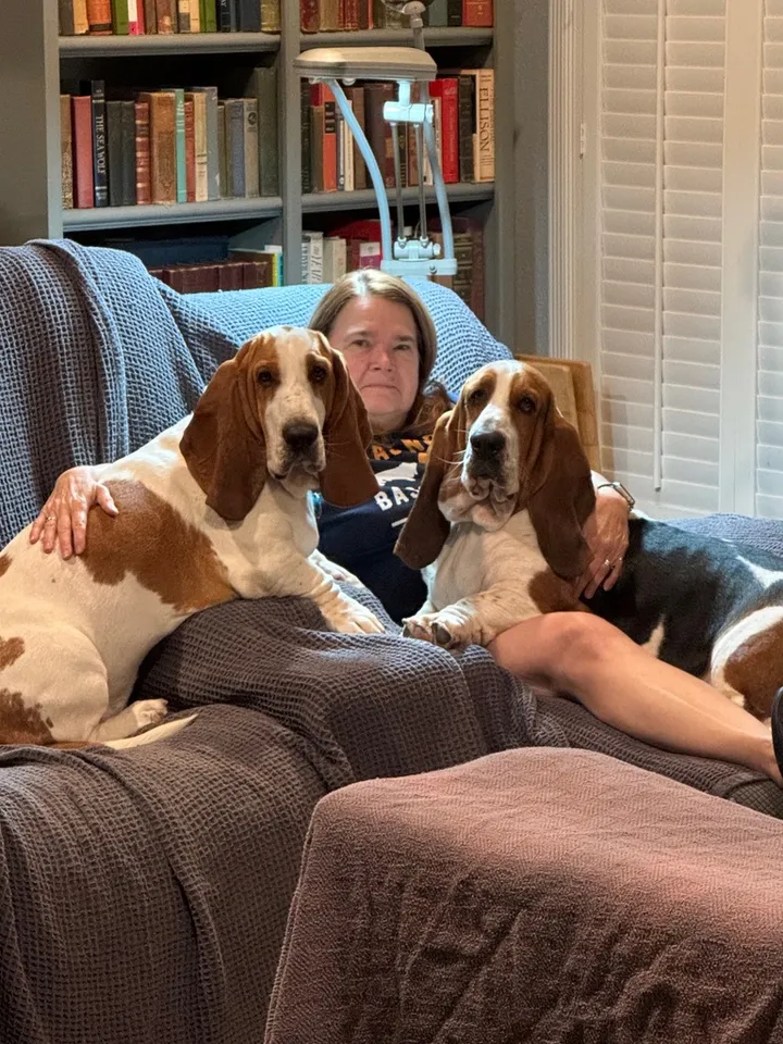 Kathi Seay at home on the couch with her two basset hounds, in front of shelves of books.